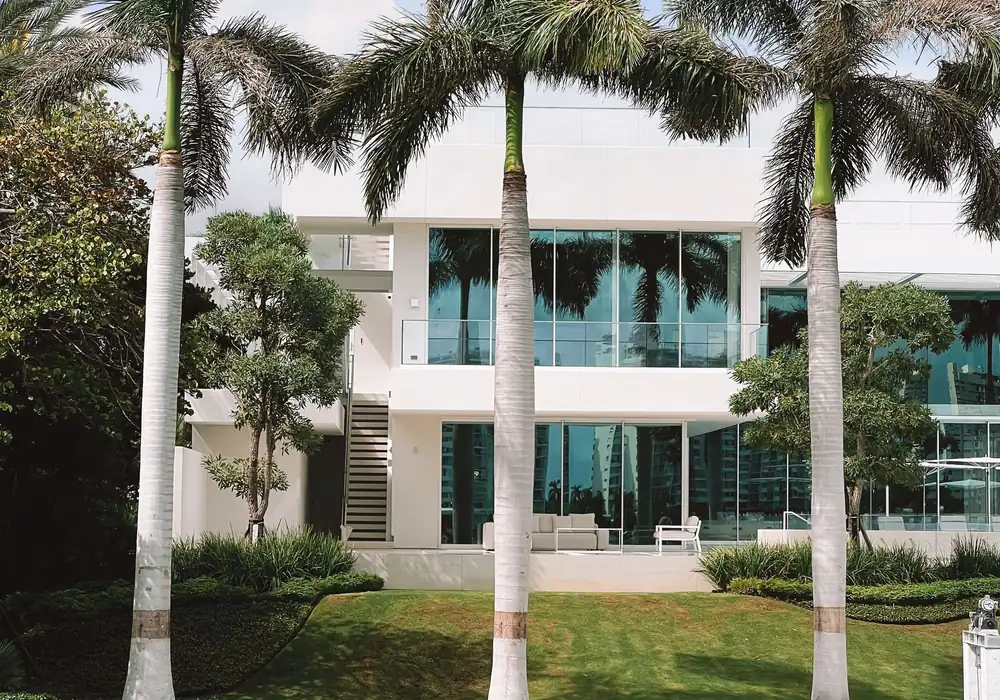 A white two-story home with floor-to-ceiling glass panels behind a lawn and tall royal palms